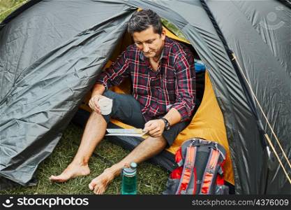 Man looking at map while planning next trip. Man relaxing in tent at camping during summer vacation. Actively spending vacations outdoors close to nature. Concept of camp life