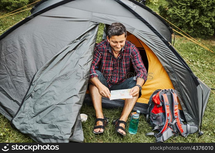 Man looking at map while planning next trip. Man relaxing in tent at camping during summer vacation. Actively spending vacations outdoors close to nature. Concept of camp life