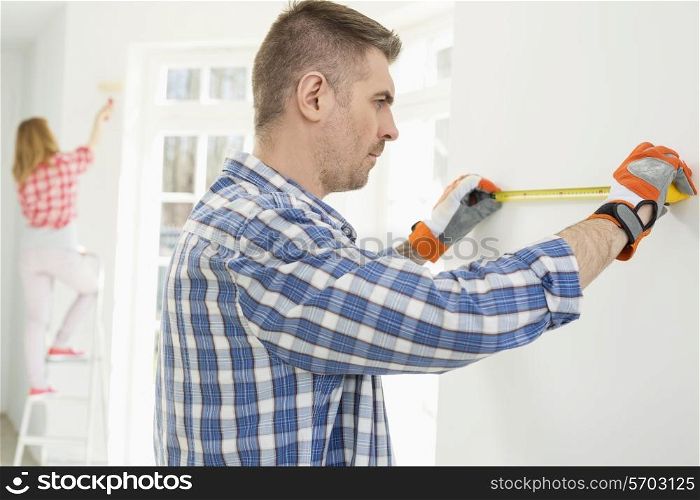 Man measuring wall with woman painting in background