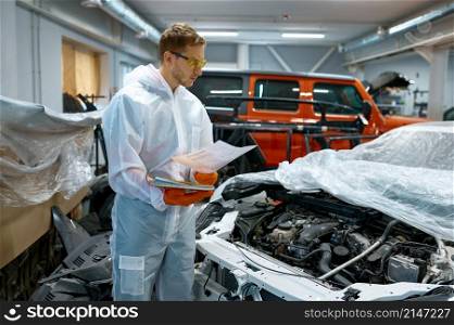 Man mechanic in safety suit looking at car engine and into paper technical certificate. Auto service. Young man mechanic looking at car engine