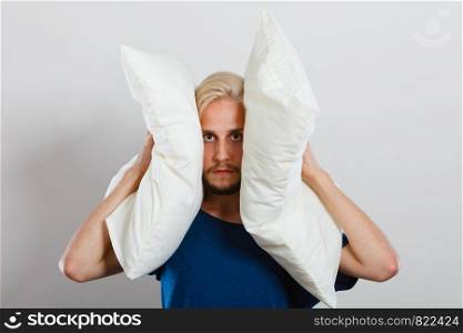 Man need silence. Sad tired young male closing ears with pillows, protecting from loud noise, have head pain. Guy not wanting to listen or can not sleep. Sad man closing ears with pillows