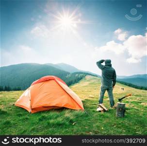 Man on mountain near orange tent looks into the distance. Man in hood stands near an orange tent and looks at the bright sun. Nearby are firewood and an ax. Summer landscape. The concept of privacy and travel.. Man on mountain near orange tent looks into the distance