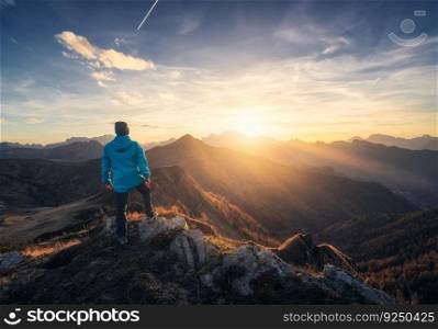 Man on stone on the hill and beautiful mountain valley in haze at colorful sunset in autumn. Dolomites, Italy. Guy, mountain ridges in fog, orange grass and trees, blue sky with sun in fall. Hiking 