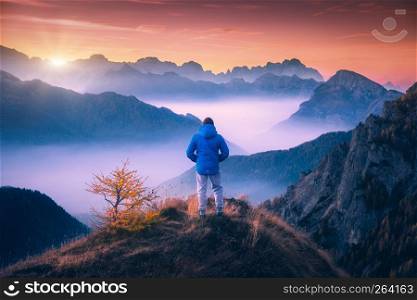 Man on the mountain peak looking on mountain valley with low clouds at colorful sunset in autumn in Dolomites. Landscape with traveler, foggy hills, forest in fall, beautiful sky at dusk in Alps