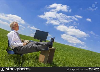 Man or male businessman relaxing feet up at a desk with a computer in a green field drinking tea or coffee