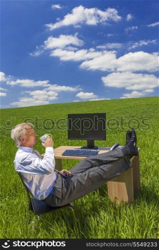 Man or male businessman relaxing feet up at a desk with a computer in a green field drinking tea or coffee