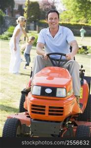 Man outdoors driving lawnmower smiling with family in background