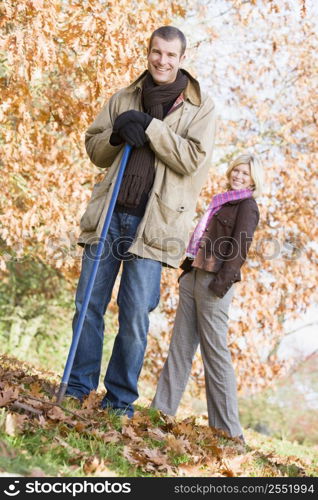 Man outdoors raking leaves and woman in background smiling (selective focus)