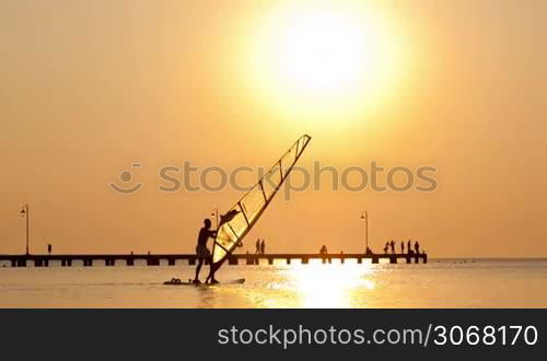 Man passing by with his windsurf or sailboard at sunset on a calm ocean against a spectacular vivid orange sky