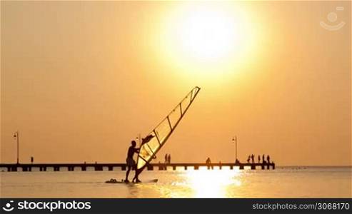 Man passing by with his windsurf or sailboard at sunset on a calm ocean against a spectacular vivid orange sky