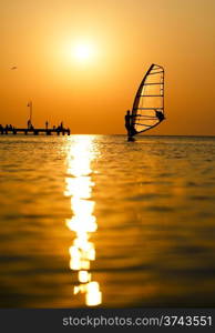 Man passing by with his windsurf or sailboard at sunset on a calm ocean against a spectacular vivid orange sky