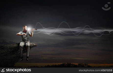 Man play violin. Young handsome man sitting on rock edge and playing violin