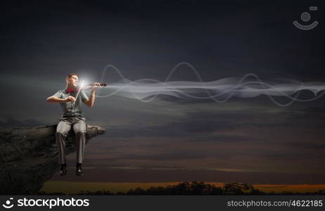 Man play violin. Young handsome man sitting on rock edge and playing violin