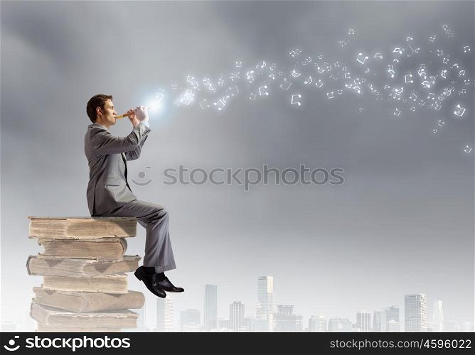 Man playing flute. Young carefree businessman sitting on pile of books and playing flute