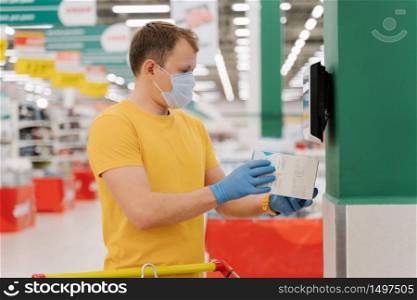 Man poses in big shopping centre, scans price of something in box, going to make purchase, wears protective medical mask and disposable rubber gloves to prevent coronavirus, uses bar code scanner