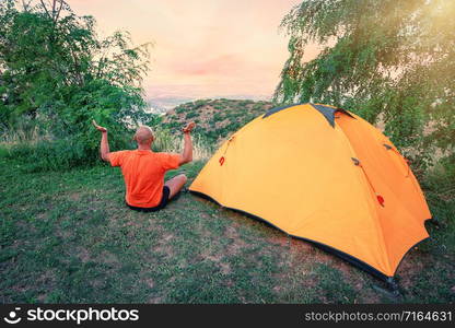 Man practices yoga near an orange tent on a hill. A man in an orange T-shirt sits in an asana near tourist tent at dawn. Concept of travel, freedom and privacy. Man practices yoga near an orange tent on hill