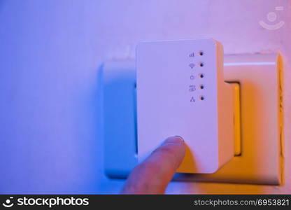 Man press with his finger on WPS button on WiFi repeater which is in electrical socket on the wall. The device help to extend wireless network in home or office.