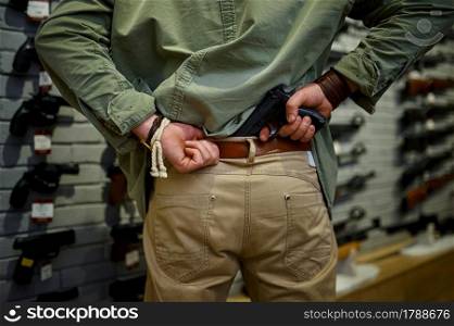 Man puts the pistol in his belt at the showcase in gun store. Weapon shop interior, ammo and ammunition assortment, firearms choice, shooting hobby and lifestyle, self protection. Man puts the pistol in his belt in gun store