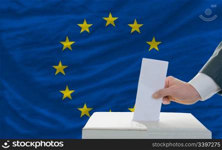 man putting ballot in a box during elections in europe in fornt of flag