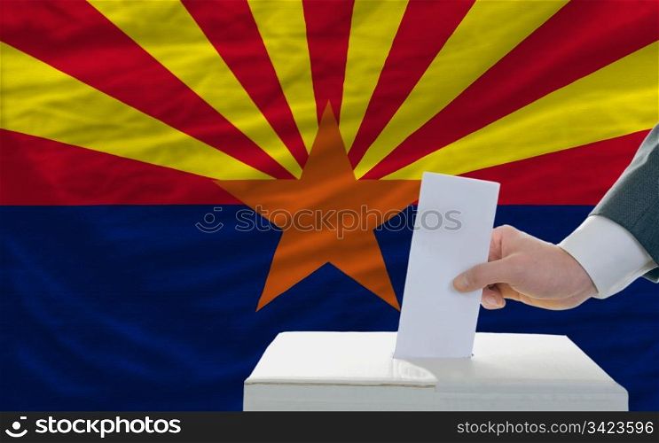 man putting ballot in a box during elections in front of flag american state of arizona