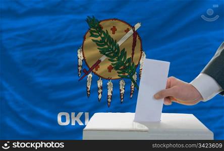 man putting ballot in a box during elections in front of flag american state of oklahoma