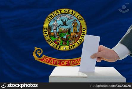 man putting ballot in a box during elections in front of flag american state of idaho