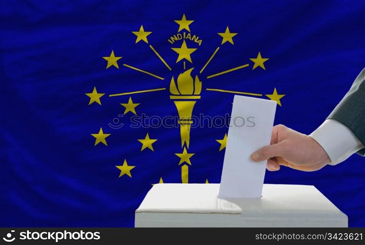 man putting ballot in a box during elections in front of flag american state of indiana