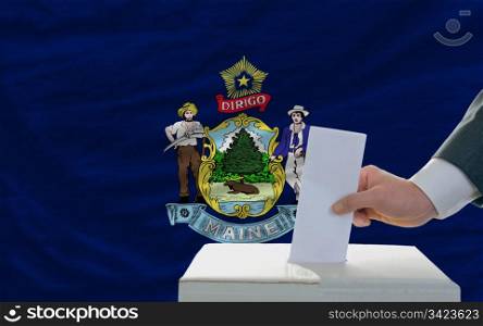 man putting ballot in a box during elections in front of flag american state of maine