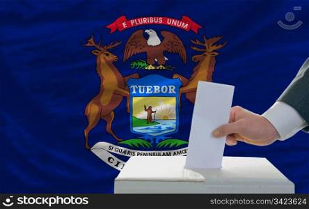 man putting ballot in a box during elections in front of flag american state of michigan