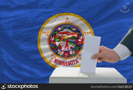 man putting ballot in a box during elections in front of flag american state of minnesota