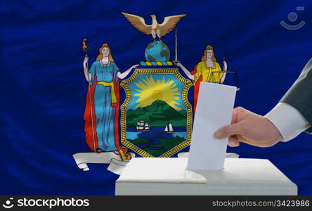 man putting ballot in a box during elections in front of flag american state of new york