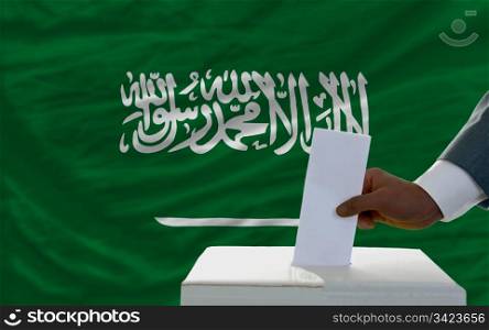 man putting ballot in a box during elections in front of national flag of saudi arabia