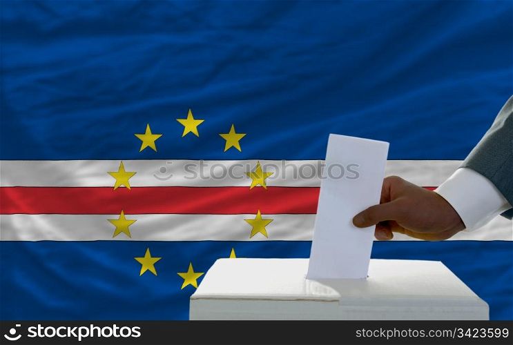 man putting ballot in a box during elections in front of national flag of capeverde