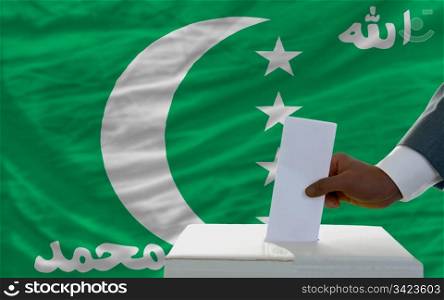 man putting ballot in a box during elections in front of national flag of comoros