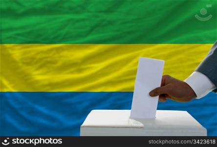 man putting ballot in a box during elections in front of national flag of gabon