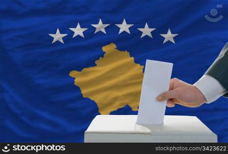 man putting ballot in a box during elections in front of national flag of kosovo