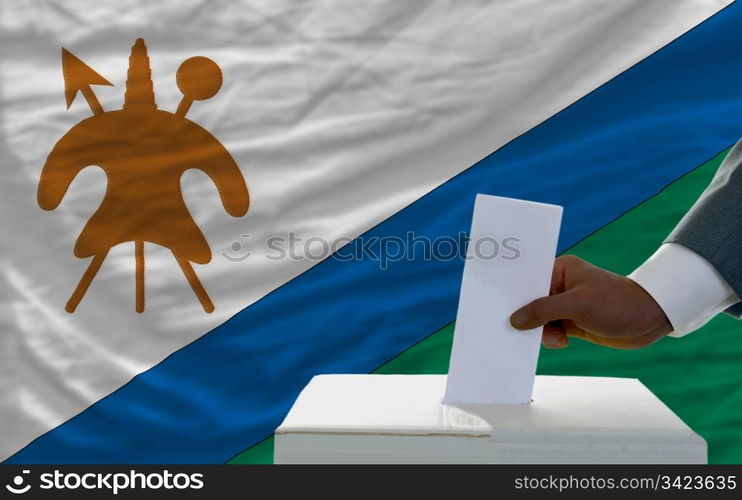 man putting ballot in a box during elections in front of national flag of lesotho