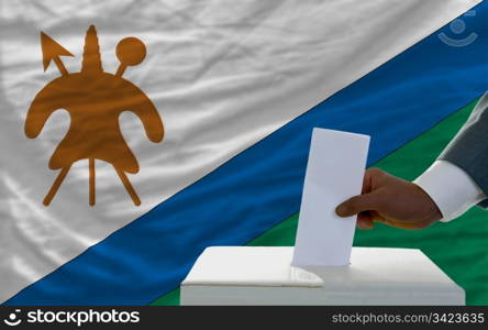 man putting ballot in a box during elections in front of national flag of lesotho