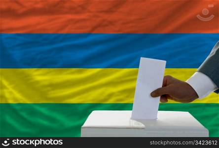 man putting ballot in a box during elections in front of national flag of mauritius