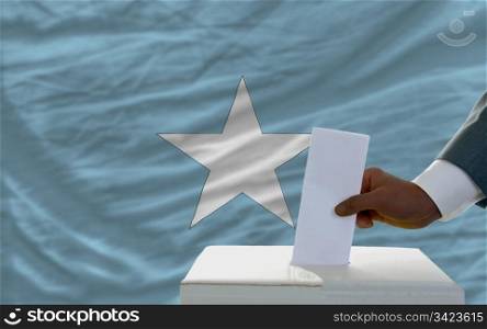 man putting ballot in a box during elections in front of national flag of somalia