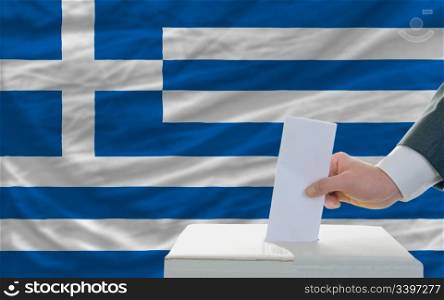 man putting ballot in a box during elections in greece in fornt of flag
