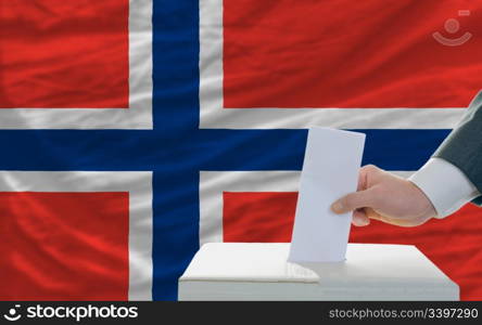 man putting ballot in a box during elections in norway in front of flag