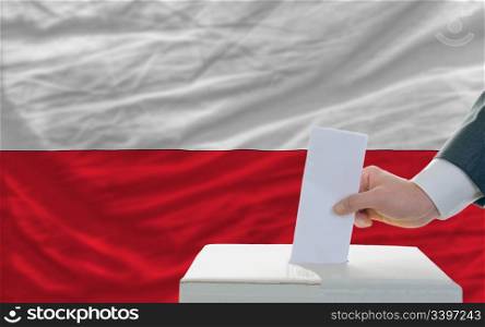 man putting ballot in a box during elections in poland in front of flag