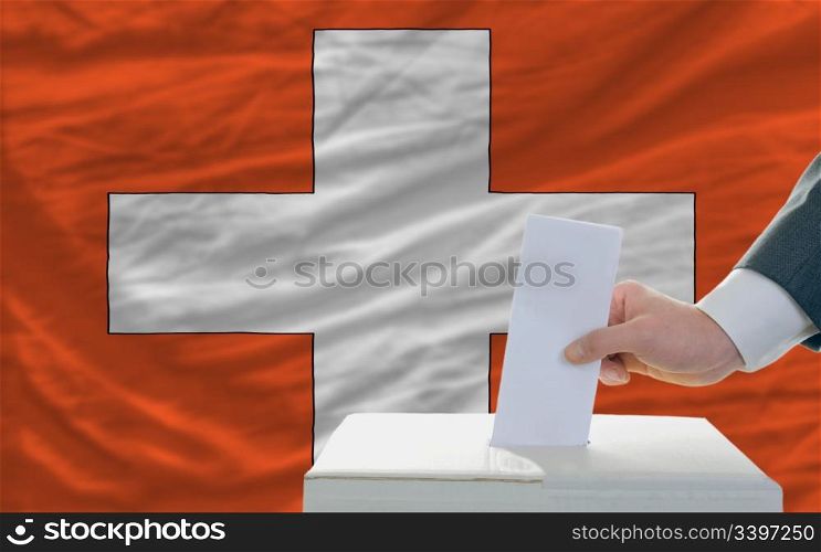 man putting ballot in a box during elections in swiss in front of flag