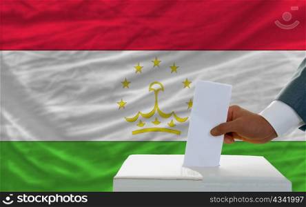 man putting ballot in a box during elections in tajikistan in front of flag