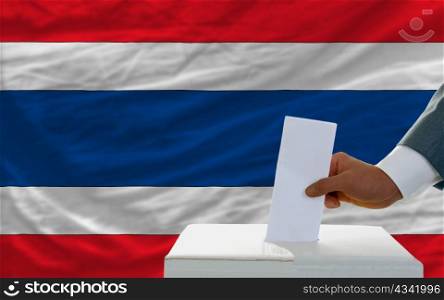 man putting ballot in a box during elections in thailand in front of flag