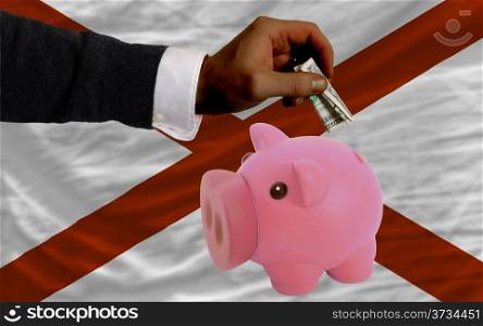 Man putting dollar into piggy rich bank flag of us state of alabama in foreign currency because of inflation