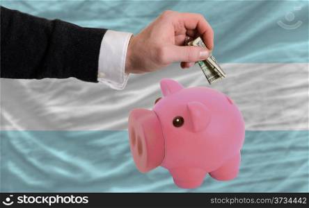 Man putting dollar into piggy rich bank national flag of argentina in foreign currency because of inflation