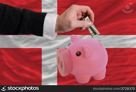 Man putting dollar into piggy rich bank national flag of denmark in foreign currency because of inflation