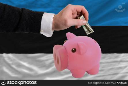 Man putting dollar into piggy rich bank national flag of estonia in foreign currency because of inflation